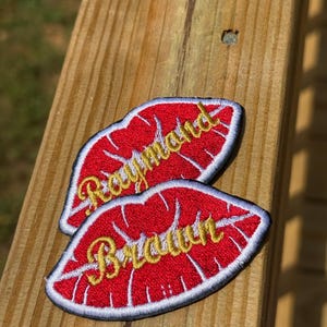 May include: Two red lip-shaped embroidered patches with gold text that reads "Raymond" and "Braun" are displayed on a wooden surface. The patches have a white border and are slightly angled.