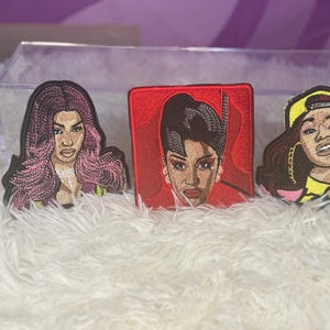 May include: Three embroidered patches featuring portraits of female figures. The patches are in the shape of the figures' faces and are in various colors, including pink, red, yellow, and black. The patches are on a white, fluffy surface.