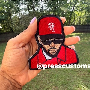 May include: Embroidered patch featuring a stylized portrait of a man wearing a red cap and sunglasses. The patch is outlined in black and has the text "@presscustoms_" at the bottom. The background is a grassy area with trees.