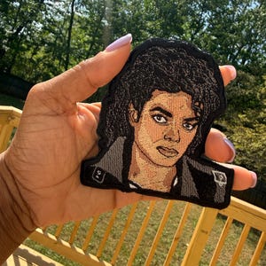 May include: Embroidered patch featuring a portrait of a person with dark curly hair and a black jacket. The patch has a black border and is held against a background of green trees and a wooden deck.