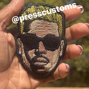May include: Embroidered patch featuring a stylized face with sunglasses. The patch has a black border, with gold-colored hair and a brown face. The text "@presscustoms_" is printed in white across the top of the patch.
