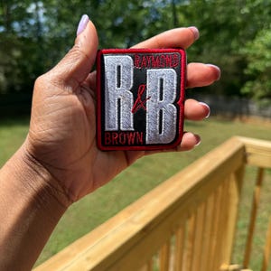 May include: Embroidered patch with the text "Raymond Brown" and the initials "R&B" in white, red, and black. The patch is rectangular with a red border. The patch is held up against a blurred green background.