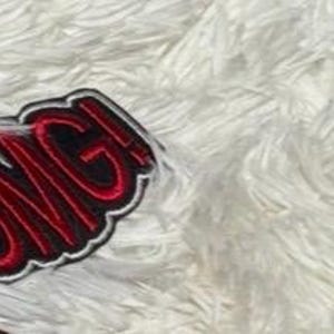 May include: A close-up of an embroidered patch with the word "OMG!" in red, outlined in black and white. The patch is on a white, fluffy surface.