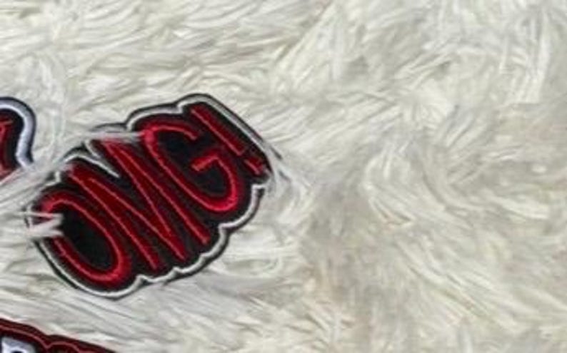 May include: A close-up of an embroidered patch with the word "OMG!" in red, outlined in black and white. The patch is on a white, fluffy surface.