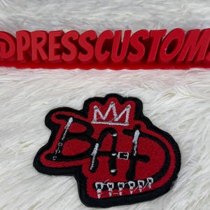 May include: Red embroidered patch with the word "BAD" in a graffiti style, topped with a crown. The patch has a black border and white accents. Above it is a red sign that reads "@PRESSCUSTOMS".