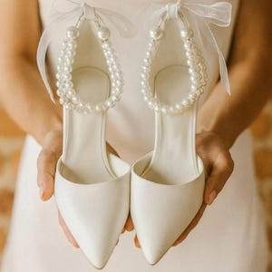 May include: A pair of white pointed-toe bridal shoes with pearl and ribbon accents. The shoes have a delicate strap of pearls that wraps around the ankle. The heels are held by a person wearing a white dress.