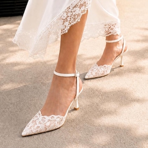 May include: A pair of white pointed-toe heels featuring intricate floral lace overlays, thin ankle straps with small buckles, and slender heels. The shoes are paired with a flowing white skirt that has a matching white lace trim at the hem.
