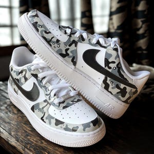 May include: Pair of white sneakers with a camouflage pattern in shades of gray and black. The sneakers feature a black swoosh logo and white laces. The soles are white with the word "AIR" printed on the side.