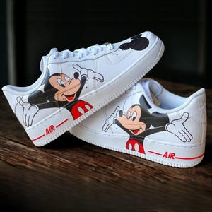 May include: White sneakers featuring a Mickey Mouse design. The shoes have a black and red Mickey Mouse graphic on the sides, with the word "AIR" in red. The soles are white with a red stripe.