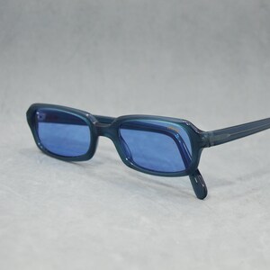 May include: Blue-tinted sunglasses with a rectangular frame. The glasses have a dark blue frame and lenses, and the arms are thin and straight. The sunglasses are set against a light grey background.