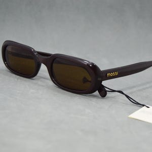 May include: Dark brown sunglasses with a rectangular frame and tinted lenses. The brand name "mossi" is printed in gold on the temple. A white tag is attached to the frame with a black string.