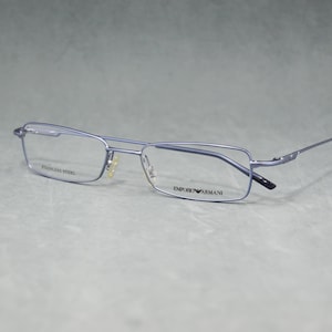 Puede incluir: Gafas rectangulares de metal azul claro. Las finas monturas llevan la marca "EMPORIO ARMANI" en la patilla derecha. La patilla izquierda dice "STAINLESS STEEL".