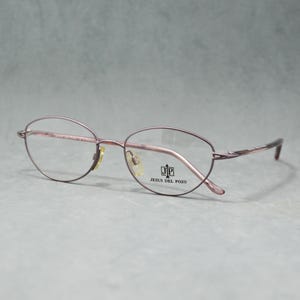 May include: Oval-shaped eyeglasses with a thin, mauve-colored metal frame. The lenses are clear, and the temples curve gently. The brand name "JESUS DEL POZO" is visible on the frame. The glasses have a classic, understated design.