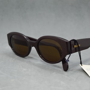 Puede incluir: Gafas de sol marrón oscuro con lentes ovaladas y montura gruesa. Las gafas de sol tienen una pequeña etiqueta adjunta con el nombre de la marca "mosssi" impreso. Las lentes son de color marrón cálido.