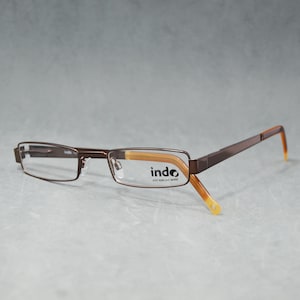 May include: Brown rectangular eyeglasses with the brand name "indo" printed on the lens. The frames are a dark brown color with light brown and orange accents on the temples. The glasses are set against a gray background.