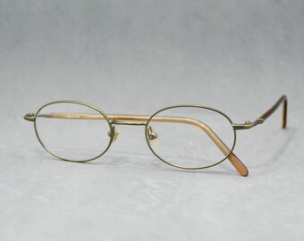 May include: Oval spectacles with a gold-coloured metal frame and clear lenses. The arms are a darker brown colour. The glasses are set against a light grey background.