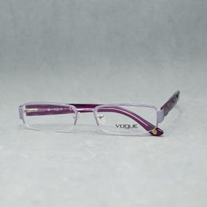 Könnte beinhalten: Violette Vogue-Brille mit rechteckigem Rahmen. Die Brille hat ein halb rahmenloses Design mit einem violetten Rahmen und Bügeln. Das Vogue-Logo ist auf dem Glas sichtbar. Die Brille ist vor einem hellgrauen Hintergrund platziert.