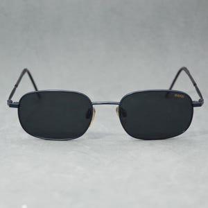 May include: Dark navy sunglasses with a rectangular frame and dark lenses. The sunglasses have thin, dark temples and a small gold-colored detail near the nose bridge. The brand name "mossi" is printed on the right lens.