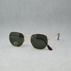 May include: Gold-framed sunglasses with dark green lenses. The glasses have an octagonal shape and a classic design. The temples are dark brown. The sunglasses are displayed on a grey surface.