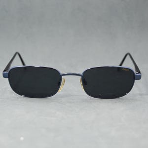 May include: Sunglasses with a dark blue frame and black lenses. The sunglasses have a rectangular shape with rounded edges. The brand name "moss" is printed on the upper left corner of the left lens. The temples are black.