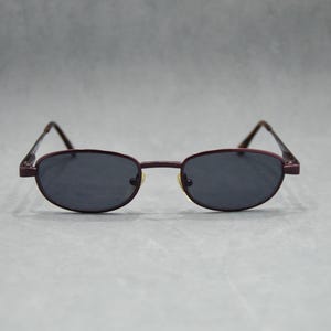 May include: Oval sunglasses with dark lenses and a burgundy metal frame. The glasses have a classic design with a thin frame and temples. The lenses are a dark grey colour, providing a sleek and stylish look.