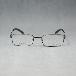 Może przedstawiać: Prostokątne okulary z ciemną metalową oprawą. Soczewki są przezroczyste, a zauszniki ciemne. Nazwy marek "BETA TITAN" i "AZZARO" są nadrukowane na soczewkach. Okulary są na neutralnym tle.