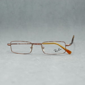 Op de afbeelding: Ray-Ban bril met een rechthoekig, bronskleurig montuur. De lenzen zijn helder en de rechterlens heeft een gele tint. De veren zijn bruin met oranje uiteinden. Het Ray-Ban logo is zichtbaar.