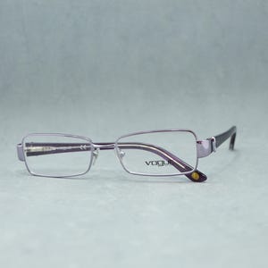 May include: A pair of Vogue eyeglasses with a rectangular frame. The frames are a light purple color with dark purple temples. The Vogue logo is visible on the lens. The glasses are set against a light gray background.
