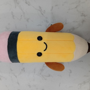 May include: A yellow pencil-shaped plush toy with a smiling face. The toy has a pink eraser, a brown tip, and small brown appendages. It features black button eyes and a black band.