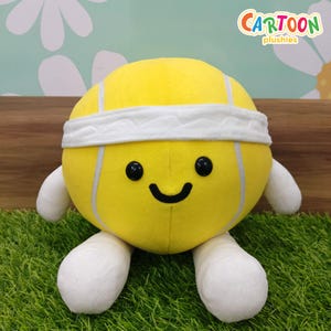 May include: A yellow tennis ball plush toy with a white headband, black eyes, and a smiling face. It has white arms and legs and is positioned on green artificial grass. The text "CARTOON plushies" is visible in the top right corner.