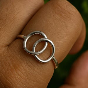 Peut inclure: Une bague en argent avec deux cercles entrelacés. La bague est en métal brillant et réfléchissant. Le design est simple et élégant, avec une esthétique minimaliste. L'arrière-plan est flou, avec des tons verts et noirs.