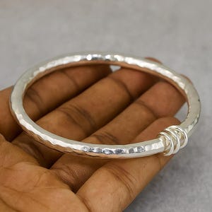 Op de afbeelding: Een zilverkleurige armband met een gehamerde textuur. De armband heeft een dik, afgerond ontwerp en een reeks opgerolde zilveren ringen op één punt. De achtergrond is effen grijs.