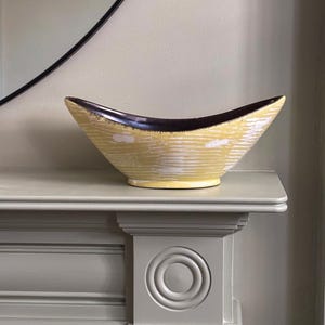 May include: A decorative ceramic bowl in a distinctive shape, with a yellow exterior and a dark brown interior. The bowl has a textured surface. It is displayed on a grey mantelpiece. The background includes a black-framed mirror.