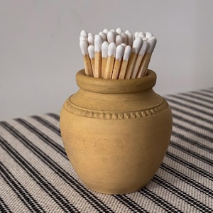 May include: A small, tan ceramic match holder filled with wooden matches. The matches have white tips and are arranged in the decorative container. The holder has a detailed, textured design around the rim. The container is likely made of ceramic.