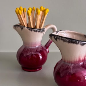 May include: Two small ceramic pitchers, one filled with matches. The pitchers are deep red with a white and black rim. The matches have yellow tips. The pitchers have a handle and are decorative.
