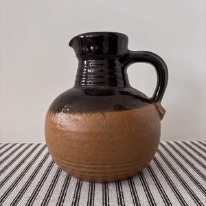May include: A brown ceramic jug with a rounded base and a dark brown top, handle, and spout. The jug has a textured surface and a classic design, suitable for serving beverages or as a decorative item.