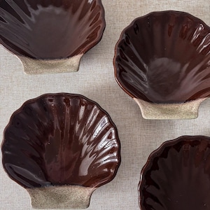 May include: Four brown ceramic shell-shaped dishes with a glossy finish. The dishes have a textured, ridged design and sit on a light beige base. The dishes are arranged on a neutral-coloured surface.