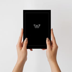 May include: A rectangular black card featuring a thin white border and a small, detailed white butterfly illustration centered in the middle. The card has a smooth, matte finish and a clean, minimalist design with high contrast between the black and white.