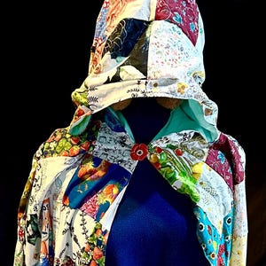 Può includere: Un mantello con cappuccio patchwork con interno blu. Il mantello presenta una varietà di motivi in tessuto colorati, tra cui disegni floreali e geometrici. Una chiusura rossa a forma di fiore fissa la parte anteriore. Il mantello è esposto su un manichino.