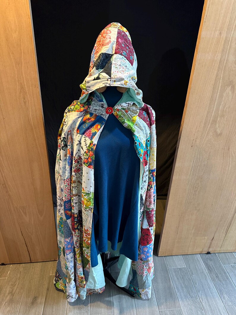 May include: A patchwork hooded cloak with a blue interior and a red button closure. The cloak features a variety of floral and patterned fabric squares in shades of red, blue, green, and white. The cloak is displayed on a mannequin.