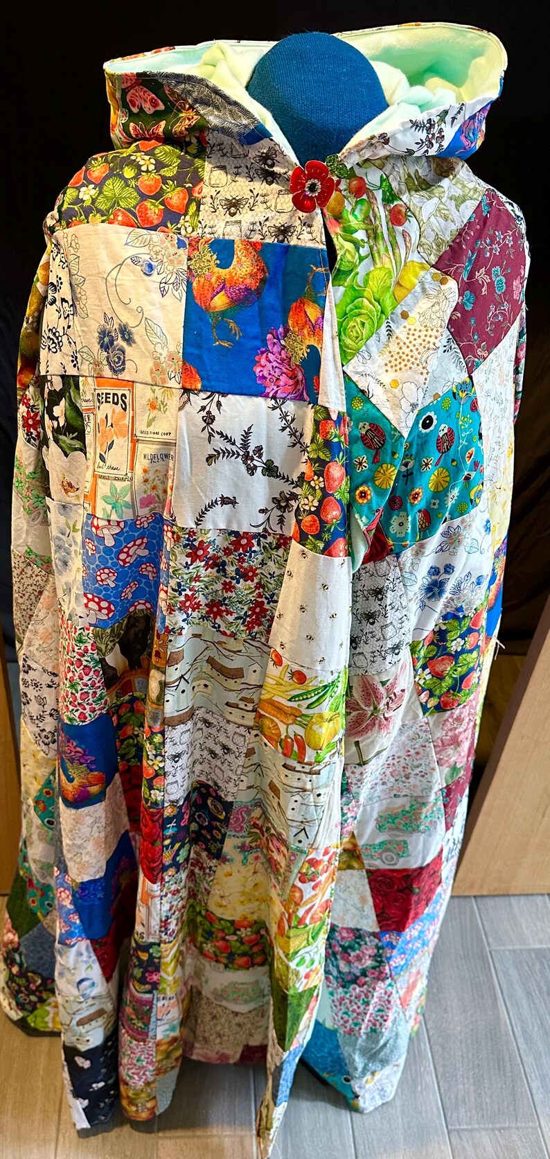 May include: A patchwork coat with a variety of fabric patterns, including floral, rooster, and botanical prints. The coat has a blue hood and a red flower closure. The coat is made of various fabric squares sewn together.