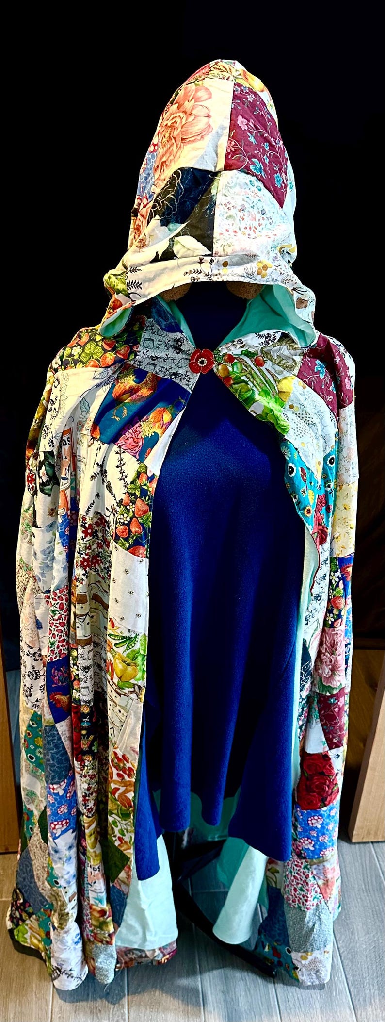 May include: A patchwork hooded cape with a blue interior. The cape features a variety of colorful fabric patterns, including floral and geometric designs. A red flower-shaped clasp secures the front. The cape is displayed on a mannequin.
