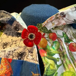 May include: A red enamel flower brooch with a black center and rhinestone accents, pinned to a patchwork fabric with bee and vegetable prints. The background is a blue knit fabric.