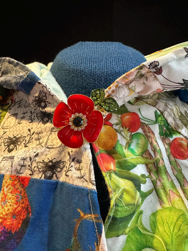 May include: A red enamel flower brooch with a black center and rhinestone accents, pinned to a patchwork fabric with bee and vegetable prints. The background is a blue knit fabric.