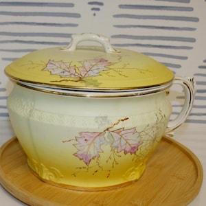 May include: A yellow ceramic chamber pot with a matching lid and a side handle. The exterior and lid feature a painted design of light purple and green leaves with thin brown stems. The rim and handle are accented with a thin gold-coloured trim.