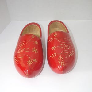 May include: A pair of red wooden clogs with a glossy finish. Each clog features a decorative floral design in a lighter color. The clogs are a traditional style, likely for decorative purposes.