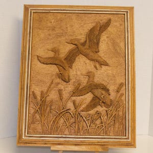 May include: A rectangular wooden wall plaque featuring a detailed relief carving of three ducks in flight over a marshy area with tall reeds and cattails. The artwork is encased in a light-coloured wooden frame with a multi-layered border design.