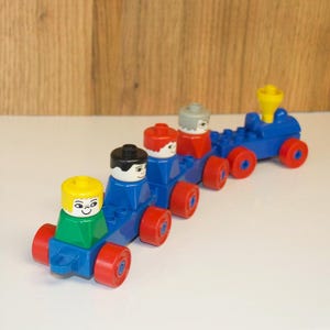 May include: A toy train constructed from interlocking plastic building blocks. The train features a blue engine with a yellow smokestack and three connected carriages, each carrying a block-shaped figure with a distinct hair colour: yellow, black, red, and grey.
