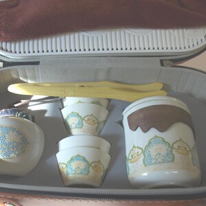 May include: A tea set inside a zipped carrying case, featuring a white ceramic teapot with blue and gold patterns, two matching small cups, a tall white canister with a dark wood lid, and light-coloured wooden tongs resting on top.