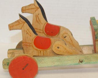 May include: A vintage wooden toy depicting two horses pulling a cart. The horses are painted light brown with black manes and red saddle details. The cart has a green base and red wheels, a classic design.